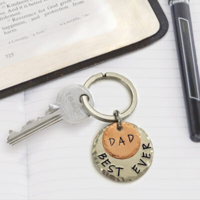 The Best Dad Ever Keychain features two metal tags—one copper engraved “DAD” and one silver with “BEST EVER”—holding a silver key, displayed on an open notebook beside a pen and a book. Read more at Stamping Cat Studio (stampingcatstudio.com).