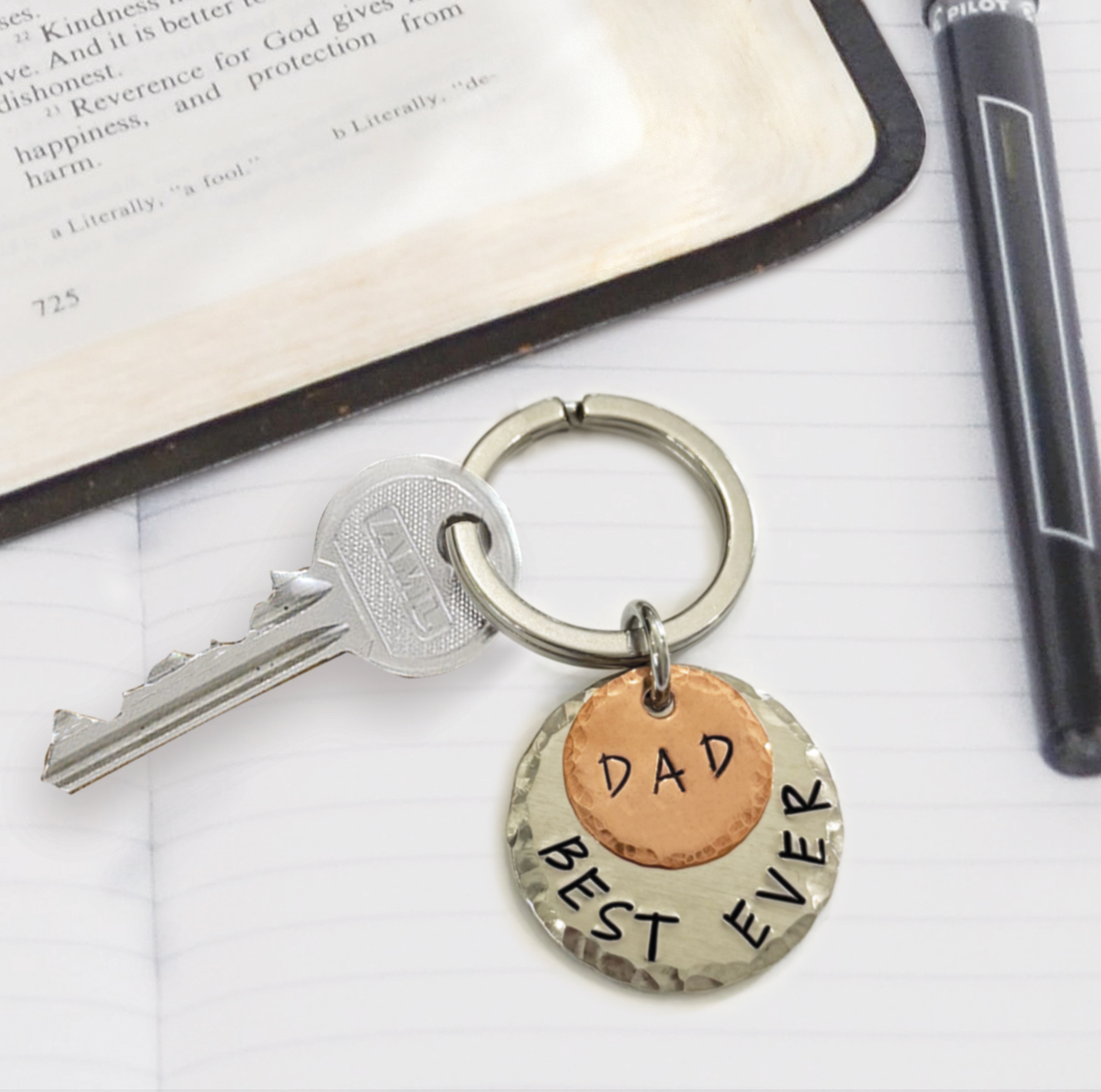 The Best Dad Ever Keychain features two metal tags—one copper engraved “DAD” and one silver with “BEST EVER”—holding a silver key, displayed on an open notebook beside a pen and a book. Read more at Stamping Cat Studio (stampingcatstudio.com).