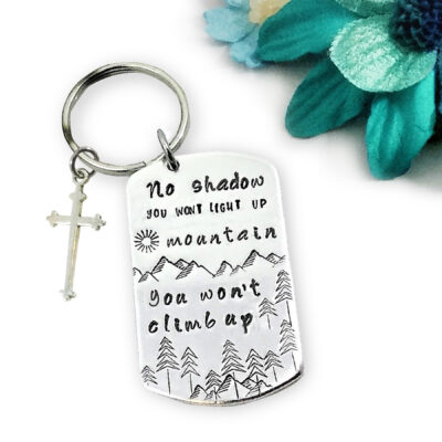 The No Shadow Key 2 is a silver keychain with a cross charm and a tag engraved with mountains, trees, and inspiring words. Blue and teal faux flowers in the background add a vibrant accent. Read more at Stamping Cat Studio (stampingcatstudio.com).