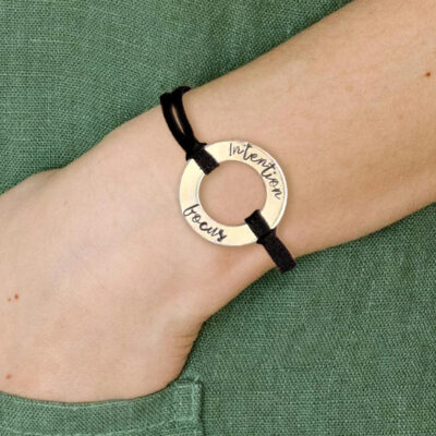 A hand wears a Men Women Personalized Washer Bracelet with a round gold-toned "Intention" and "focus" charm on a black cord. A green textured sleeve appears in the background. Read more at Stamping Cat Studio (stampingcatstudio.com).