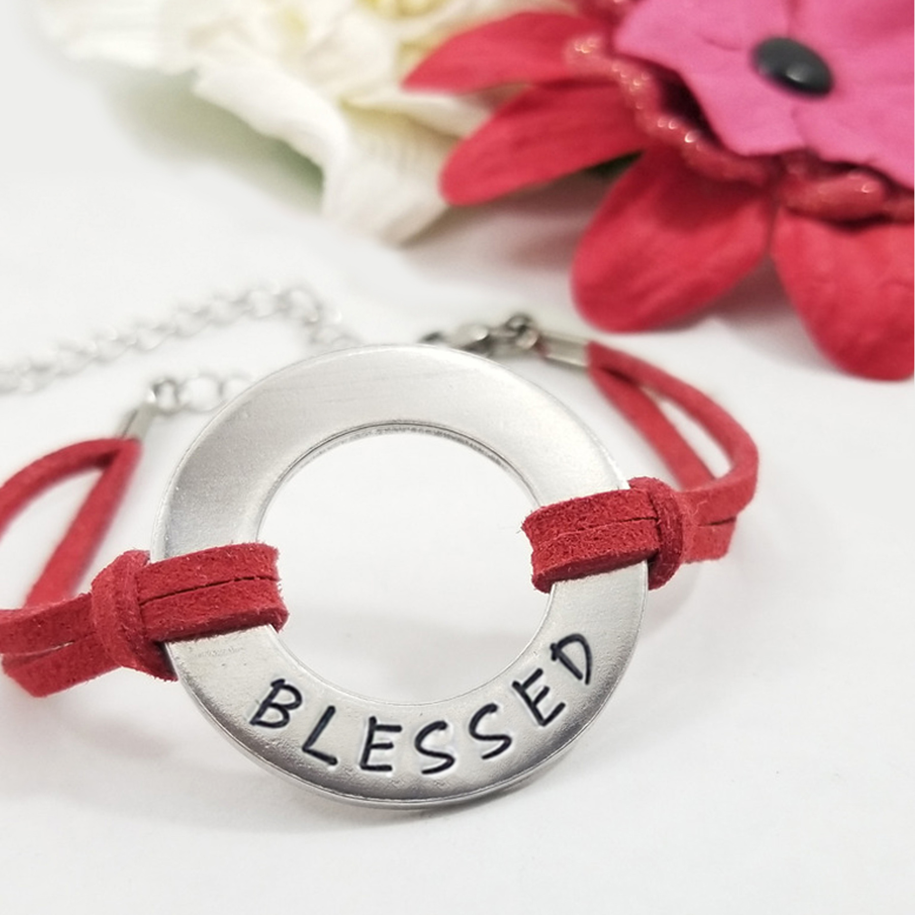 A silver circular charm engraved with "BLESSED" accents the Men Women Personalized Washer Bracelet in red suede, displayed on a white surface with blurred red and white fabric flowers in the background. Read more at Stamping Cat Studio (stampingcatstudio.com).
