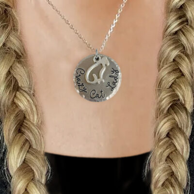 A person with two blonde braids models the Crazy Cat Lady Necklace—a silver pendant with a cat outline and “Crafty Cat Lady” text—against a blurred background that highlights the necklace and their neck. Read more at Stamping Cat Studio (stampingcatstudio.com).
