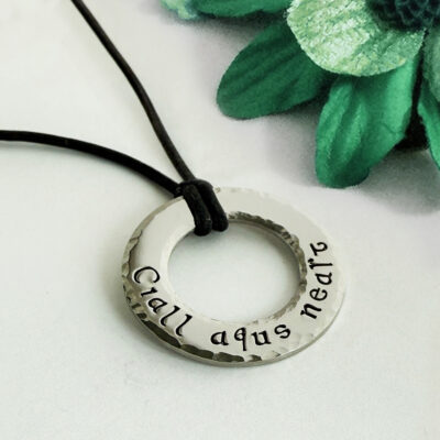 The Irish quote washer is a silver circular pendant engraved with "Ciall agus neart," hanging from a black cord, shown resting on a light surface next to a green artificial flower with textured petals. Read more at Stamping Cat Studio (stampingcatstudio.com).