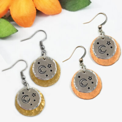 The Man In The Moon ear main earrings feature two overlapping hammered metal discs—one silver with engraved moons and stars, and one gold or copper beneath—with fish hook wires, shown near orange and green artificial flower petals. Read more at Stamping Cat Studio (stampingcatstudio.com).
