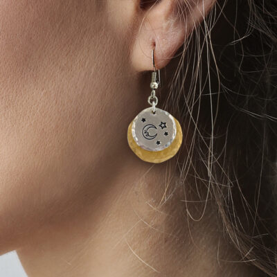 Close-up of a person's ear wearing the Man In The Moon nugold ear—a double-layered dangle earring featuring a silver disc engraved with stars and a crescent moon, layered over a textured gold disc. Brown hair partially frames the ear and earring. Read more at Stamping Cat Studio (stampingcatstudio.com).