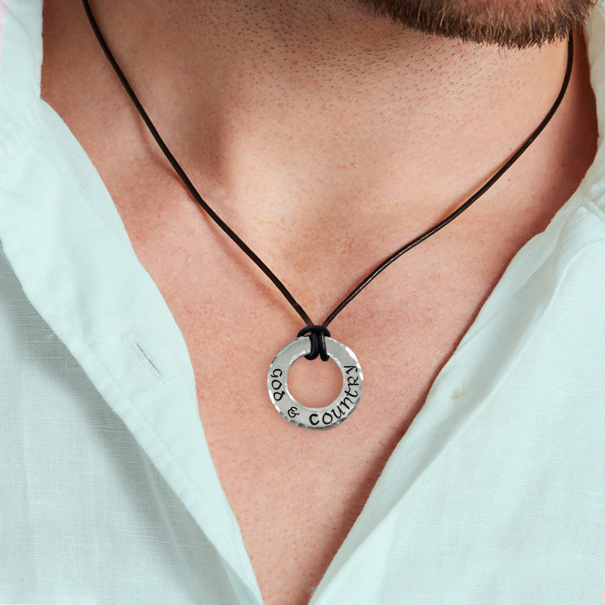 A person wearing a light green collared shirt with the top button open displays the "Pers washer neck man 2" necklace—a round silver pendant engraved with “GOD & COUNTRY” on a black cord. Only their lower face and neck are visible. Read more at Stamping Cat Studio (stampingcatstudio.com).