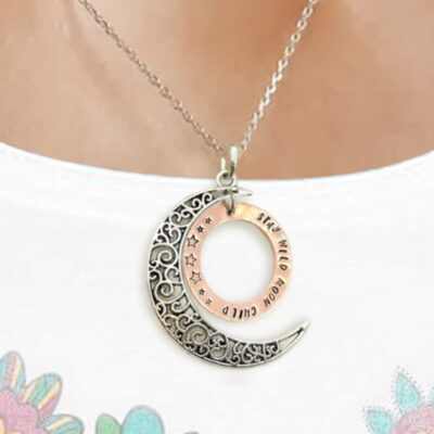 A Stay Wild Moon Child Copper Circle Necklace with a silver crescent moon and bronze engraved "I LOVE YOU TO THE MOON AND BACK" pendant, featuring stars and swirling patterns, is worn over a white floral shirt. Read more at Stamping Cat Studio (stampingcatstudio.com).