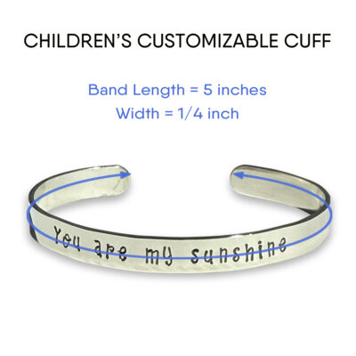 The Children’s Customizable Cuff Bracelet is silver with “You are my sunshine” engraved, a slightly open end, and measures 5" x 1/4". Blue lines and arrows show dimensions. Add your custom text above the engraving. Read more at Stamping Cat Studio (stampingcatstudio.com).