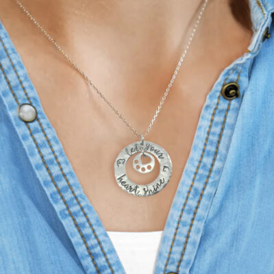 A person wears a light blue denim shirt and the Heart Shine neck 1, a silver necklace with a round pendant featuring a cut-out paw print and the engraving "Let your heart shine"—a perfect gift for animal lovers. Read more at Stamping Cat Studio (stampingcatstudio.com).