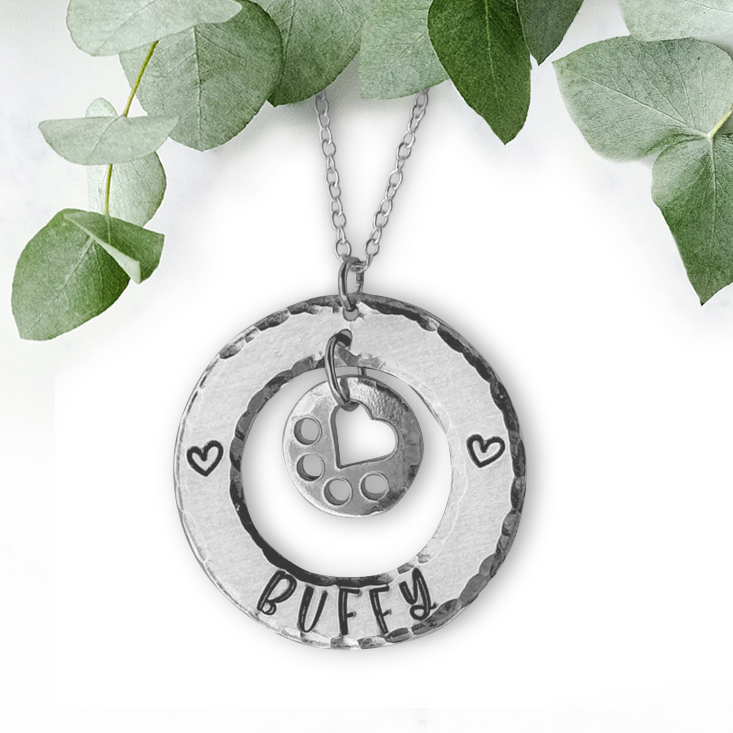 The Heart Shine neck Buffy necklace features two silver circular pendants: the larger is engraved with “BUFFY” and two hearts, while the smaller has a cut-out heart with dotted details, shown against a white background and green leaves. Read more at Stamping Cat Studio (stampingcatstudio.com).