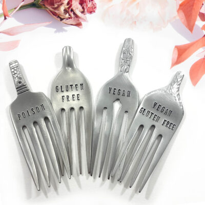 The Charming Custom Cheese Marker Forks set features four upright forks stamped "POISON," "GLUTEN FREE," "VEGAN," and "VEGAN GLUTEN FREE." Pink flowers and leaves on a white background add an elegant, whimsical touch. Read more at Stamping Cat Studio (stampingcatstudio.com).
