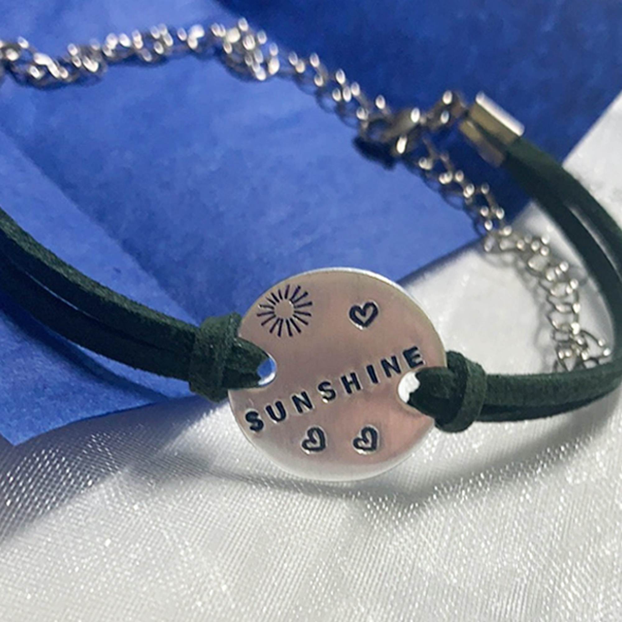 A close-up of the Charming Boho Ankle Bracelet shows its round silver "SUNSHINE" charm with a sun design and three hearts on a dark green cord, displayed against white and blue textured fabrics. Read more at Stamping Cat Studio (stampingcatstudio.com).