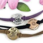 Three Charming Boho Ankle Bracelets: silver with a cat on a crescent moon (purple cord), copper "YOU’RE MY SUNSHINE" with sun (black cord), and gold with butterfly and fairy (brown cord). Pink petals above. Read more at Stamping Cat Studio (stampingcatstudio.com).