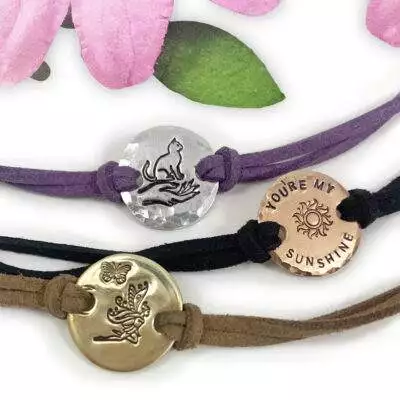 Three Charming Boho Ankle Bracelets: silver with a cat on a crescent moon (purple cord), copper "YOU’RE MY SUNSHINE" with sun (black cord), and gold with butterfly and fairy (brown cord). Pink petals above. Read more at Stamping Cat Studio (stampingcatstudio.com).