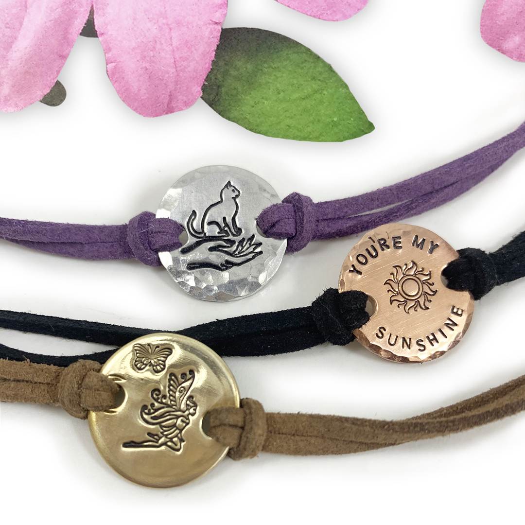 Three Charming Boho Ankle Bracelets: silver with a cat on a crescent moon (purple cord), copper "YOU’RE MY SUNSHINE" with sun (black cord), and gold with butterfly and fairy (brown cord). Pink petals above. Read more at Stamping Cat Studio (stampingcatstudio.com).