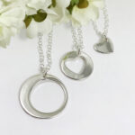 Three necklaces from the Always Family Generations Necklace Set—featuring a large circle, a heart cutout circle, and a small solid heart—hang on silver chains, displayed against white artificial flowers on a white background. Read more at Stamping Cat Studio (stampingcatstudio.com).