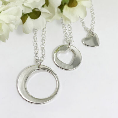 Three necklaces from the Always Family Generations Necklace Set—featuring a large circle, a heart cutout circle, and a small solid heart—hang on silver chains, displayed against white artificial flowers on a white background. Read more at Stamping Cat Studio (stampingcatstudio.com).