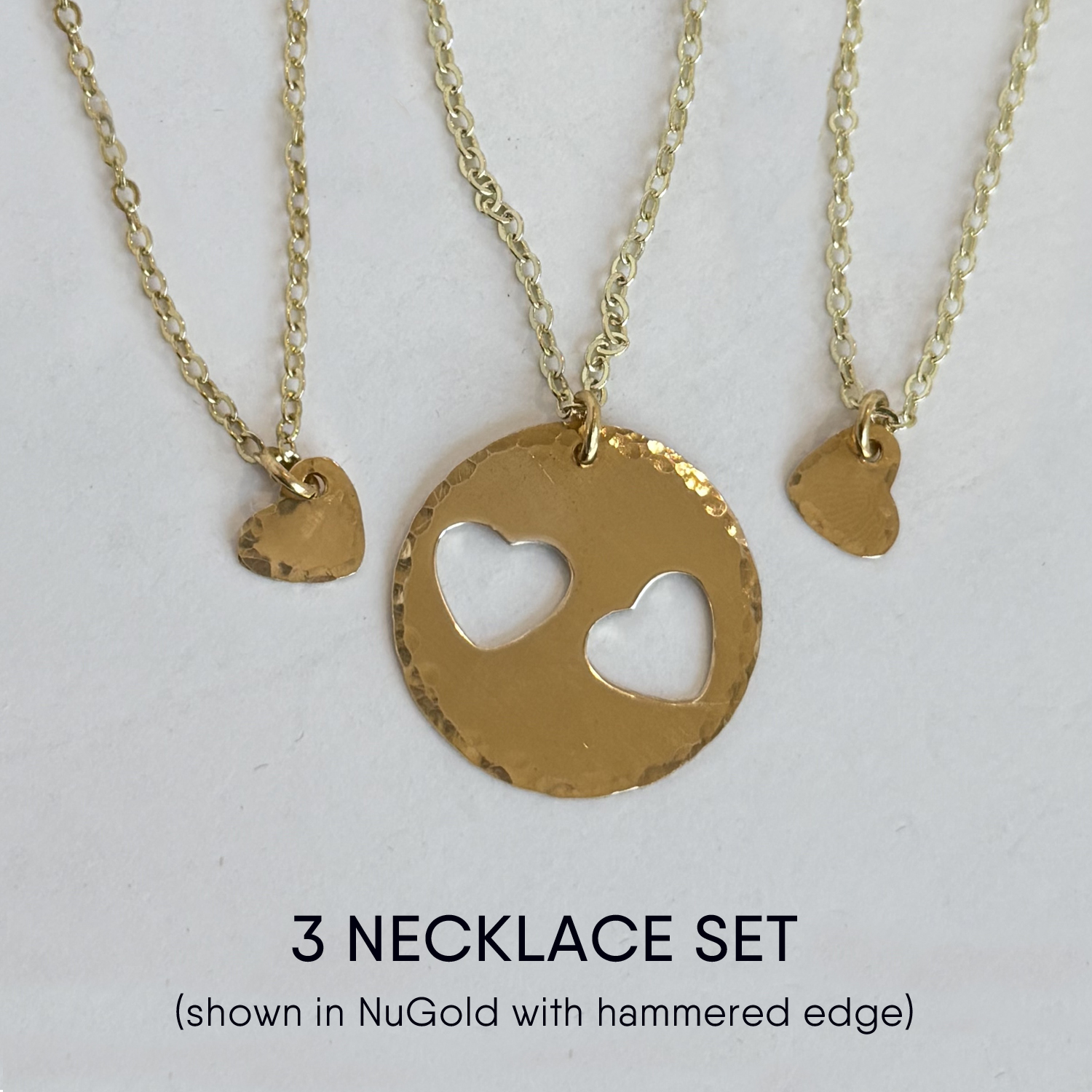 The Mother Daughter Heart Necklace Set includes three gold-toned necklaces: two with small hammered heart pendants and one with a larger hammered disc featuring two heart-shaped cutouts, all on delicate chains. Read more at Stamping Cat Studio (stampingcatstudio.com).