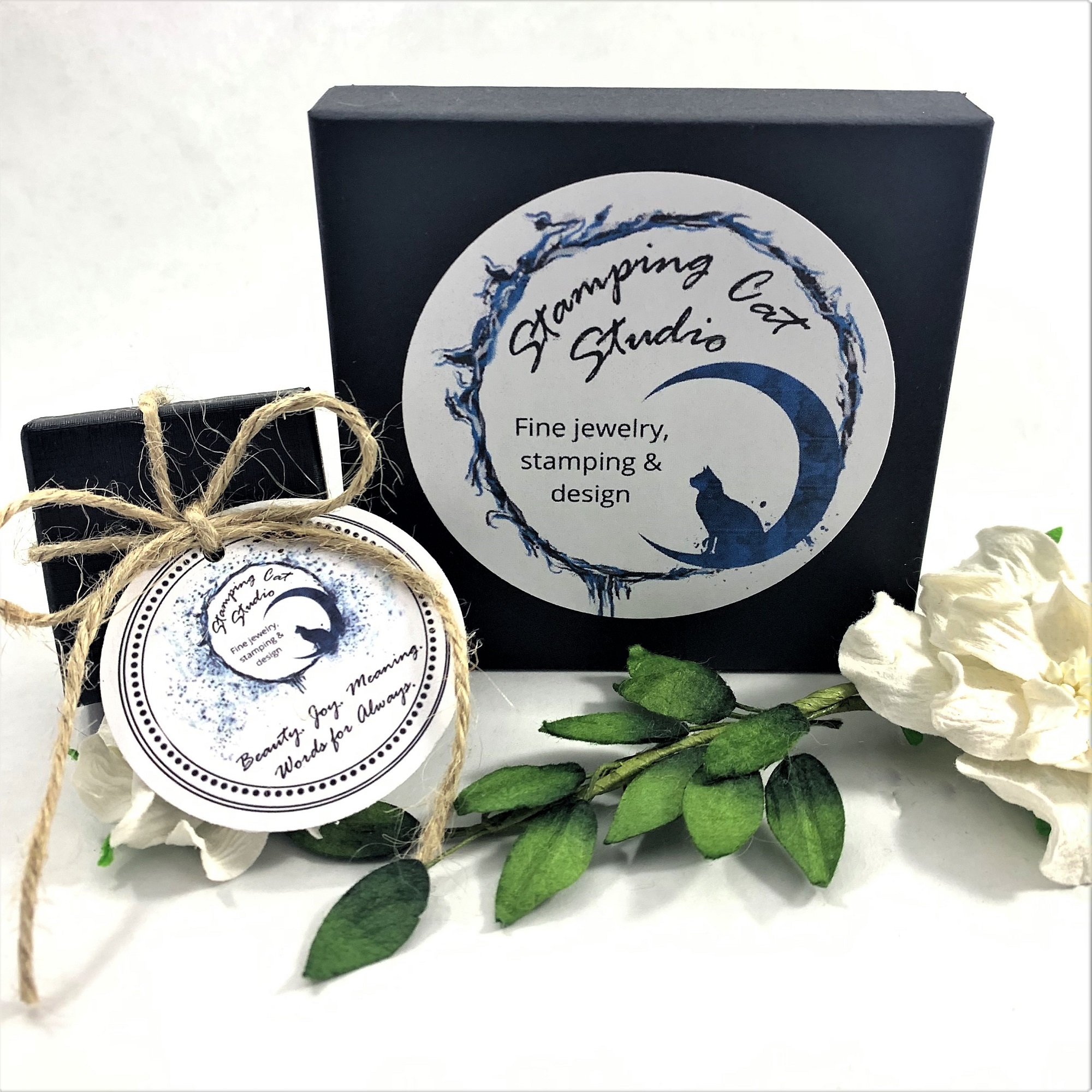 A black gift box with a circular “Stamping Cat Studio” label sits beside a twine-tied box, surrounded by white artificial flowers and greenery, showcasing the Vintage Flatware Bracelet with Heart Charm. Read more at Stamping Cat Studio (stampingcatstudio.com).