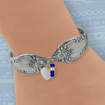 The Vintage Flatware Bracelet with Heart Charm, silver-toned with a floral pattern, features a dangling metallic heart and blue-clear beads, stylishly worn on the wrist against a blue geometric background. Read more at Stamping Cat Studio (stampingcatstudio.com).