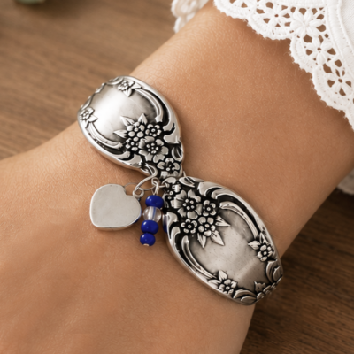 A Vintage flatware lifestyle bracelet made from two curved, floral-embossed spoon handles on a wrist, featuring a small silver heart charm and two blue beads dangling from the center. Read more at Stamping Cat Studio (stampingcatstudio.com).