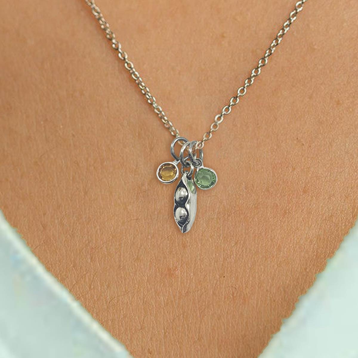 The Peas In A Pod Best Friend Necklace is a silver chain with a small pea pod pendant holding two peas and two round gemstone charms, one yellow and one light green—stunning on bare skin and a light blue shirt. Read more at Stamping Cat Studio (stampingcatstudio.com).