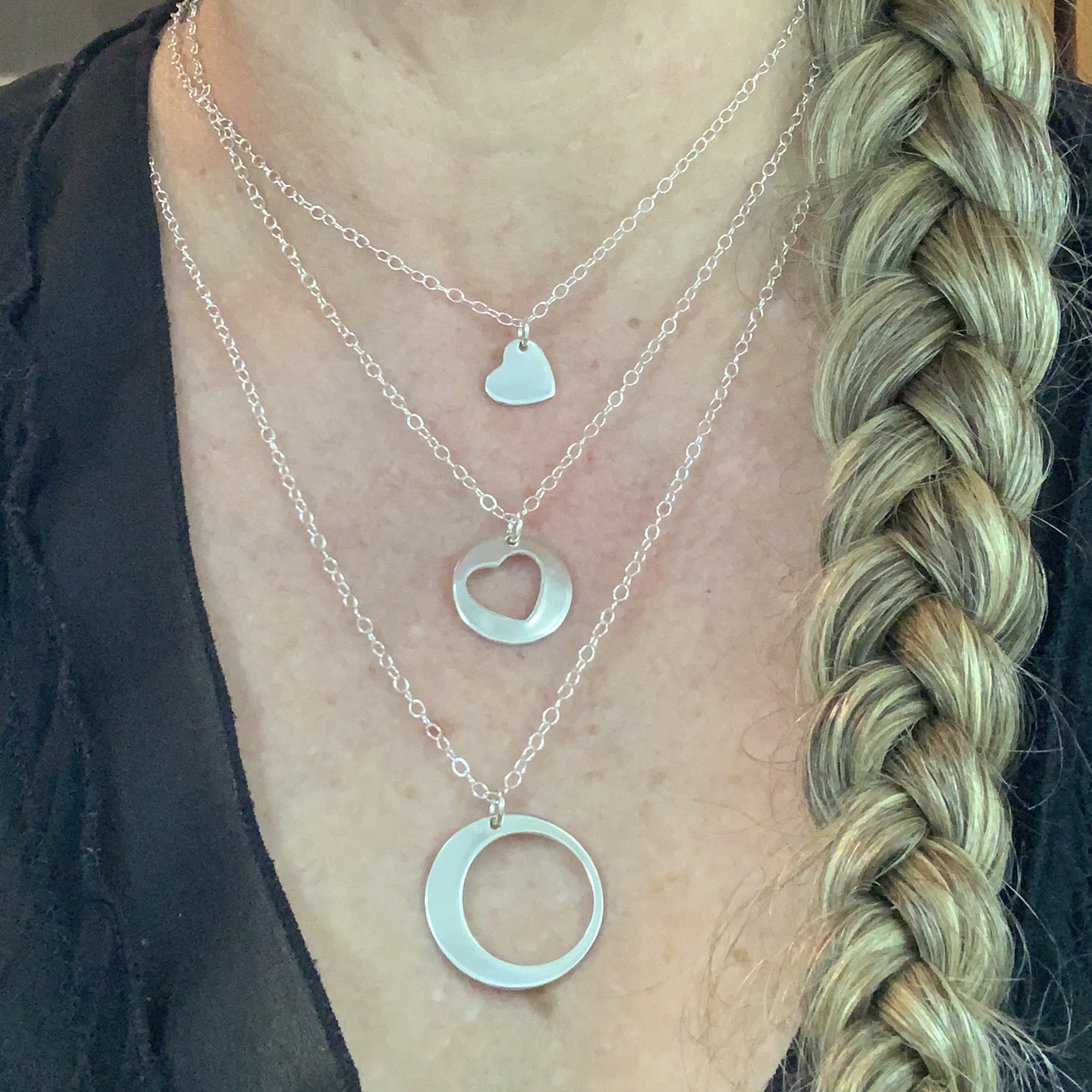 A person with a blond braid, dressed in a black top, wears the All My Heart Necklace Set: three silver necklaces featuring a small heart pendant, a circle pendant with heart cutout, and a long necklace with an open circle. Read more at Stamping Cat Studio (stampingcatstudio.com).