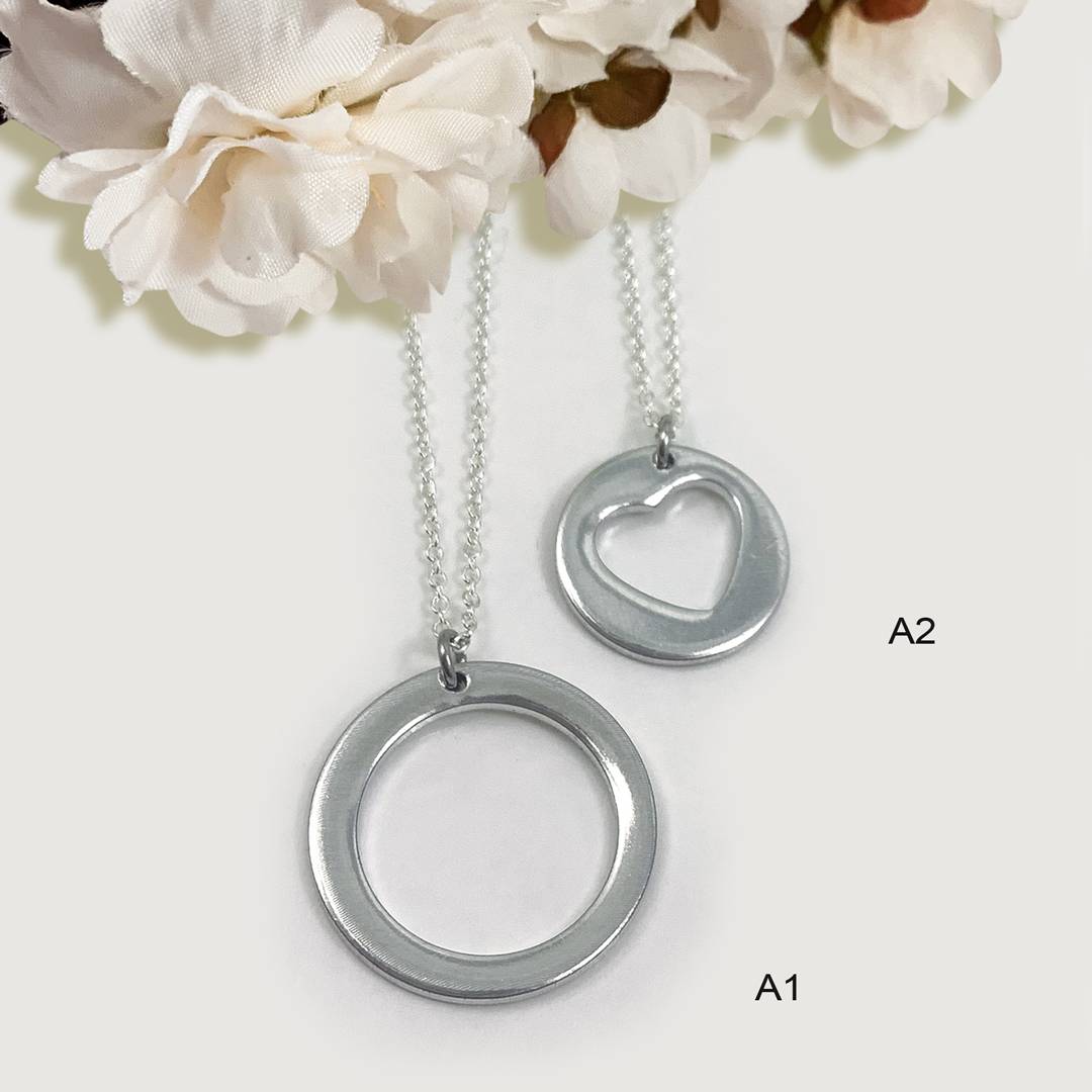 Two silver pendants from the All My Heart Necklace Set hang on chains: Pendant A1 is a plain circle, and Pendant A2 features a heart-shaped cutout. Both are displayed against white silk flowers with "A1" and "A2" labels on a white background. Read more at Stamping Cat Studio (stampingcatstudio.com).