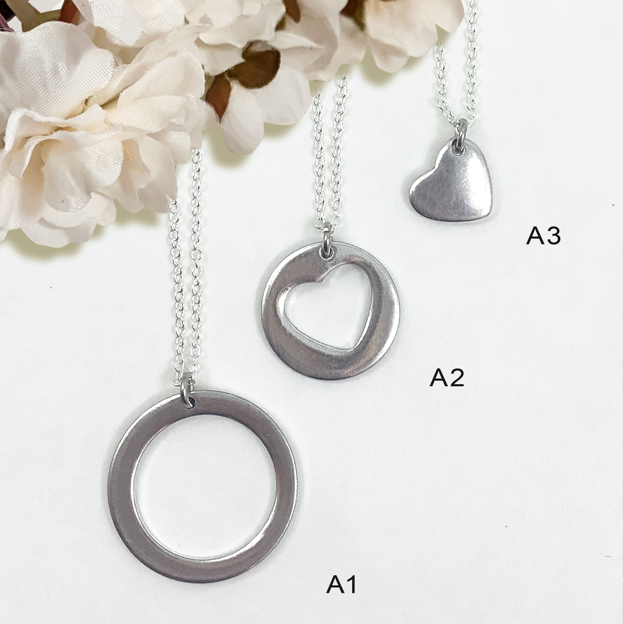 Three necklaces from the All My Heart Necklace Set are displayed vertically on silver chains against a white background with cream flowers: a circle pendant, a circle with cut-out heart, and a solid heart pendant. Read more at Stamping Cat Studio (stampingcatstudio.com).