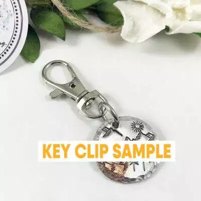 A round metal key clip engraved with mountains, trees, and stars features “KEY CLIP SAMPLE” in bold. Green leaves, a white flower, and a Baby Feet and Heart Necklace are partially visible in the background. Read more at Stamping Cat Studio (stampingcatstudio.com).