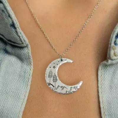 The Crescent Moon Dreamy Mountain Scene Necklace, featuring engraved mountains, trees, and stars, rests on the wearer's skin as it hangs from a delicate chain beneath a partially open light blue denim shirt. Read more at Stamping Cat Studio (stampingcatstudio.com).