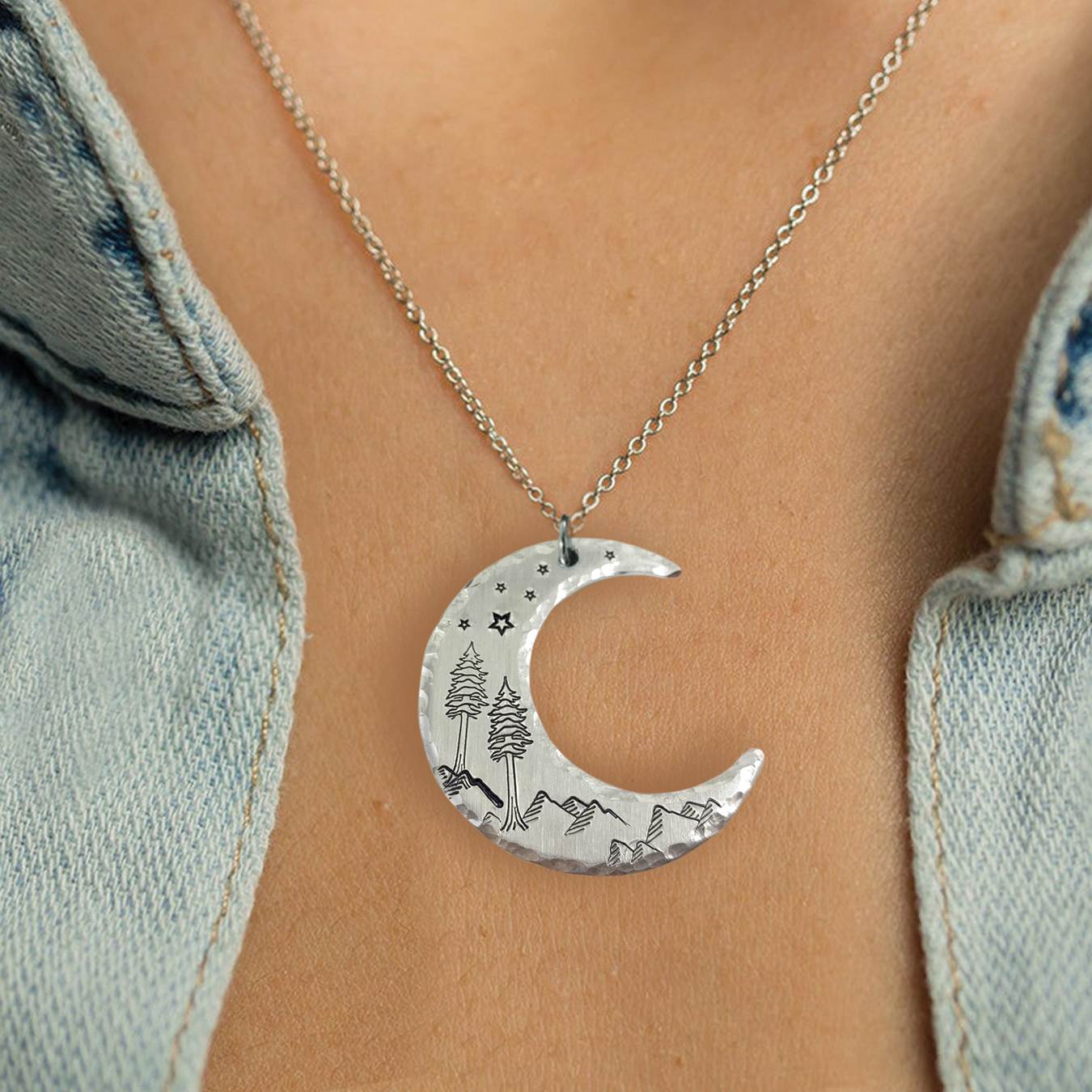 The Crescent Moon Dreamy Mountain Scene Necklace, featuring engraved mountains, trees, and stars, rests on the wearer's skin as it hangs from a delicate chain beneath a partially open light blue denim shirt. Read more at Stamping Cat Studio (stampingcatstudio.com).