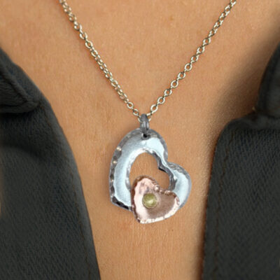 A close-up of the Mixed Metal Heart Necklace, showcasing two overlapping hearts—one large, hammered silver heart and a smaller copper-tone heart with a center gem—worn against light skin and a dark, open shirt collar. Read more at Stamping Cat Studio (stampingcatstudio.com).