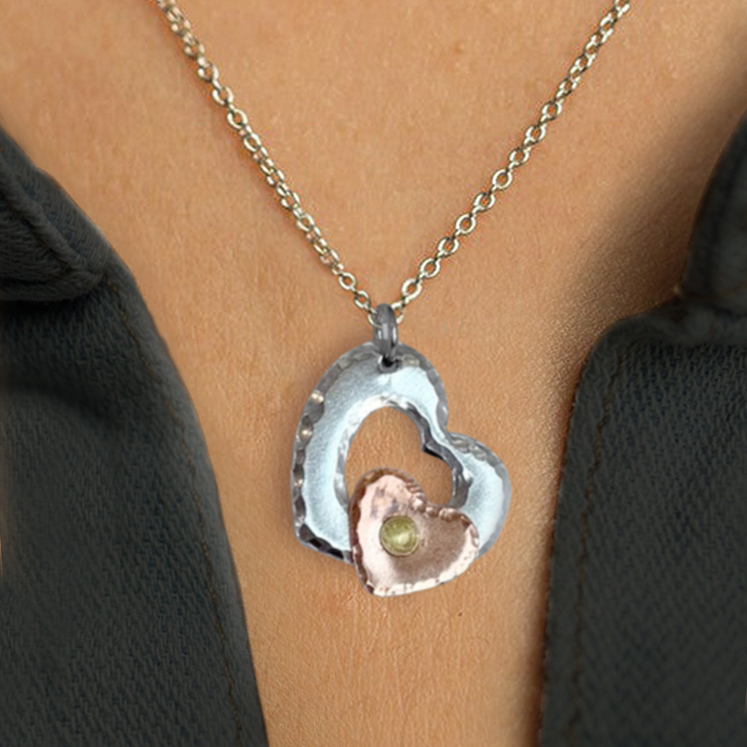 A close-up of the Mixed Metal Heart Necklace, showcasing two overlapping hearts—one large, hammered silver heart and a smaller copper-tone heart with a center gem—worn against light skin and a dark, open shirt collar. Read more at Stamping Cat Studio (stampingcatstudio.com).
