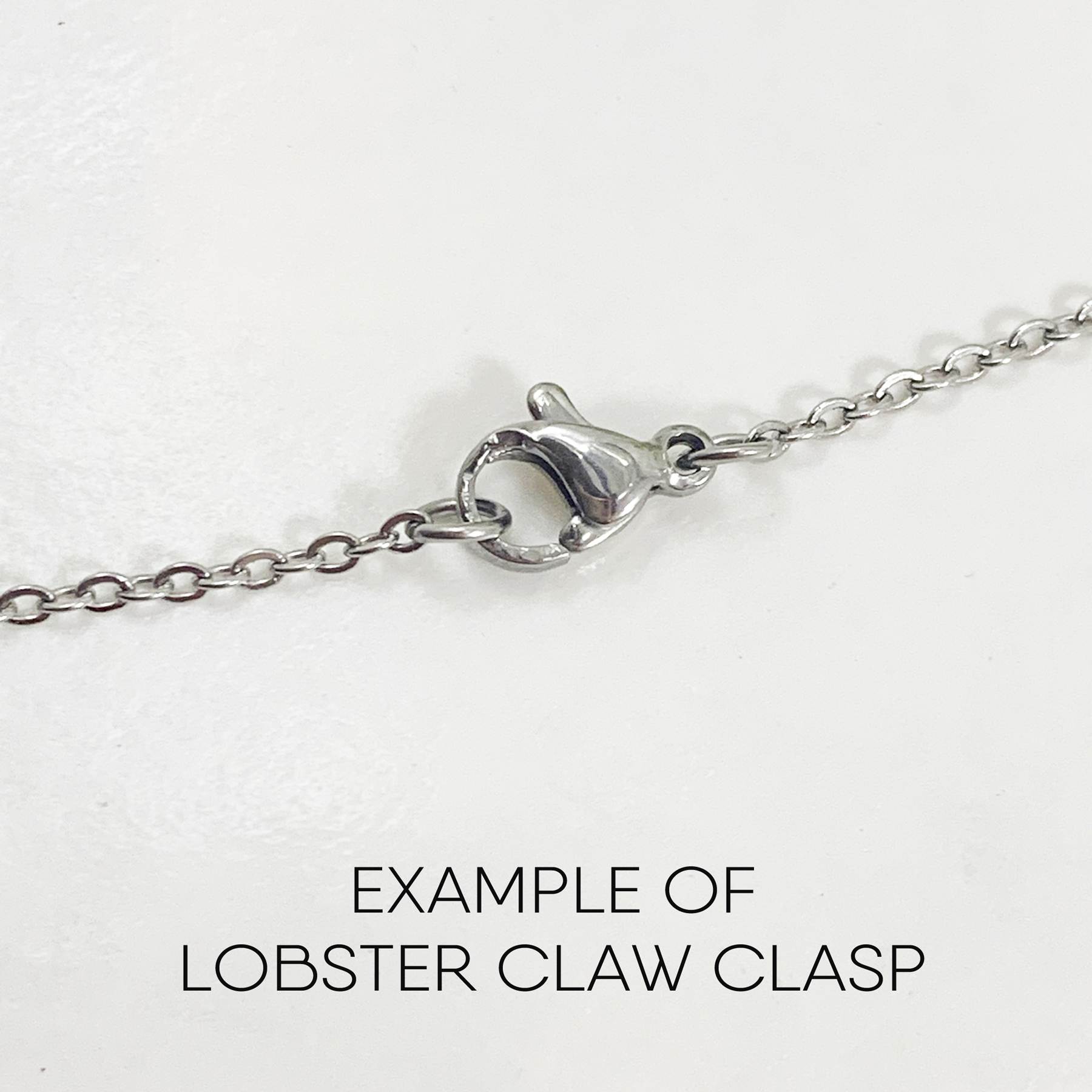 A close-up of the Dreamy Sun, Moon And Stars Necklace features an oval lobster claw clasp with a small lever in the center. Below the chain, “EXAMPLE OF LOBSTER CLAW CLASP” is written on a white background. Read more at Stamping Cat Studio (stampingcatstudio.com).