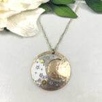 The Dreamy Sun, Moon And Stars Necklace features a silver and gold crescent moon pendant with star cutouts and gold accents, hanging from a twisted silver chain. It’s shown resting on a white surface beside green leaves and a white flower. Read more at Stamping Cat Studio (stampingcatstudio.com).