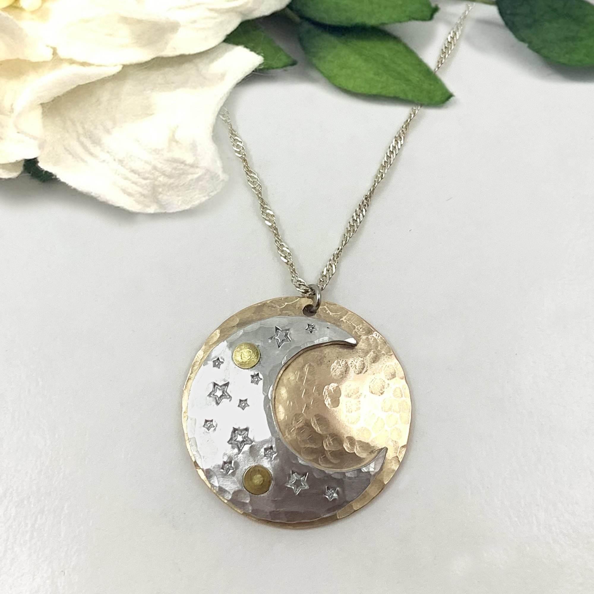 The Dreamy Sun, Moon And Stars Necklace features a silver and gold crescent moon pendant with star cutouts and gold accents, hanging from a twisted silver chain. It’s shown resting on a white surface beside green leaves and a white flower. Read more at Stamping Cat Studio (stampingcatstudio.com).