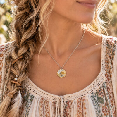 A person with a braided hairstyle wears a Dreamy Sun Moon Stars liifestyle 2 silver necklace, featuring a round pendant with a gold crescent moon and small star cutouts against a floral patterned top. Read more at Stamping Cat Studio (stampingcatstudio.com).