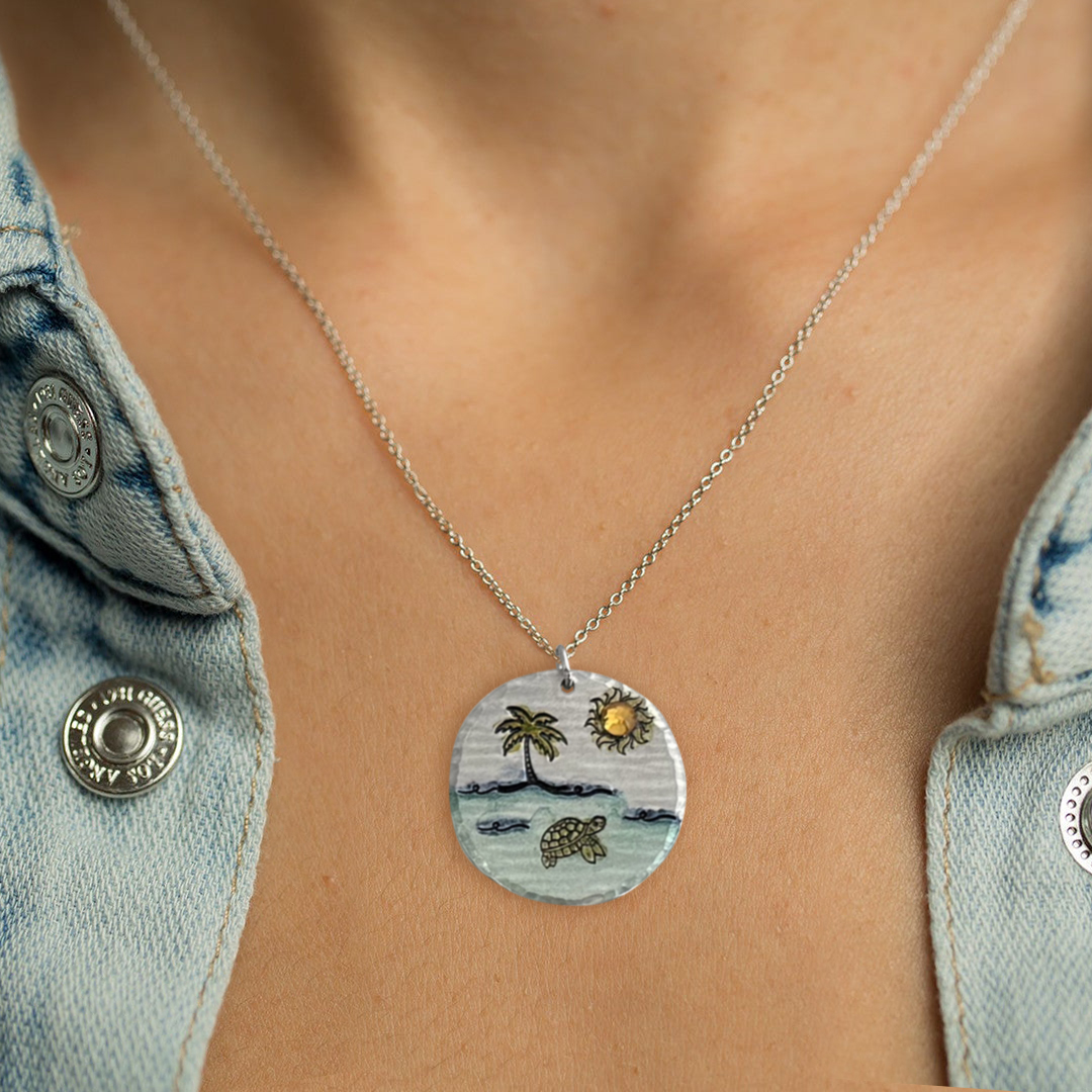 A close-up of someone wearing the Dreamy Beach Scene crystal, a round pendant with a palm tree, sun, and turtle on a beach, paired with a light blue denim shirt with metal buttons and an open collar. Read more at Stamping Cat Studio (stampingcatstudio.com).