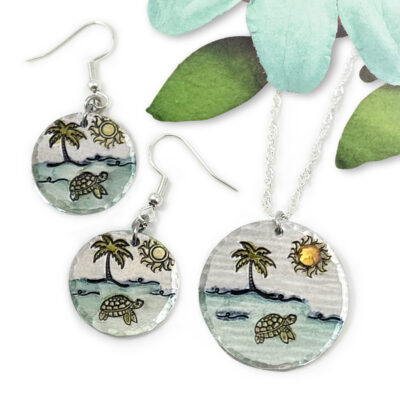 Dreamy Beach Scene set main includes round earrings and a necklace pendant with hand-painted turtle, palm tree, sun, waves, and sand designs. Displayed with a green paper leaf and blue flower for decoration. Read more at Stamping Cat Studio (stampingcatstudio.com).