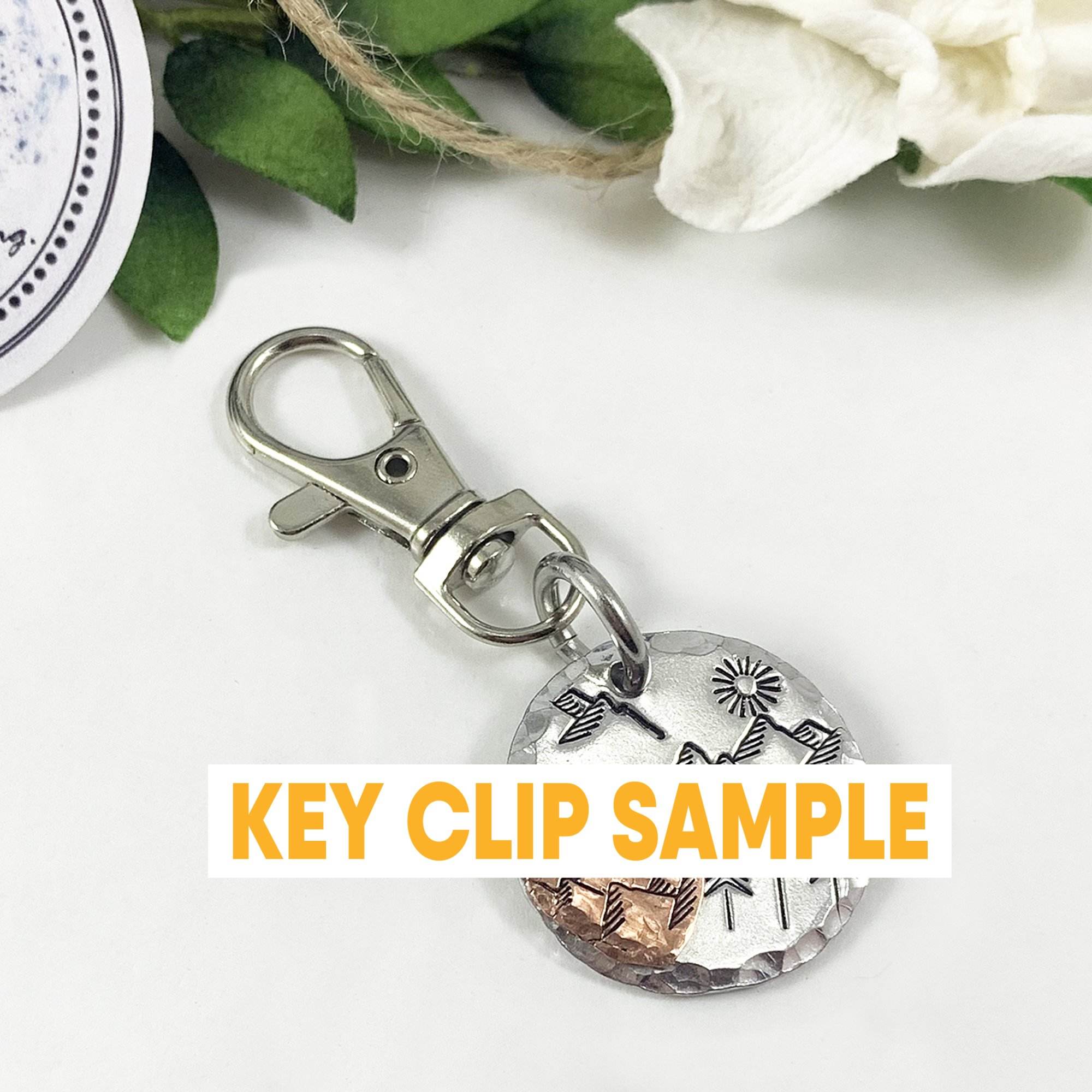 The "My Love Key Ring" is a metal key clip with a round pendant engraved with mountains, trees, and sun. A bold yellow and white “KEY CLIP SAMPLE” label covers the center. It’s shown among green leaves and a white flower in the top right. Read more at Stamping Cat Studio (stampingcatstudio.com).