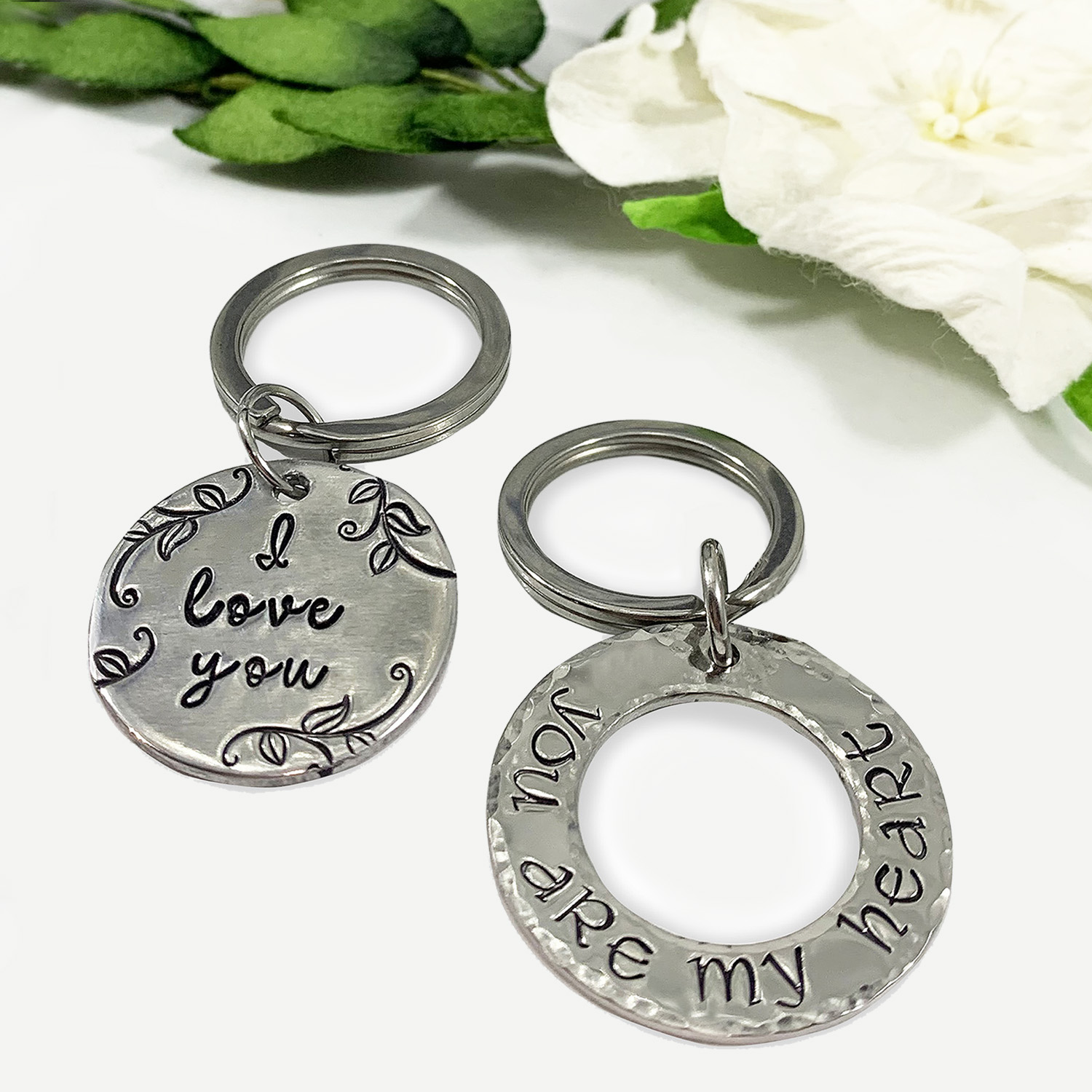 The My Love key ring main, a round silver keychain engraved with "I love you" and leafy designs, lies on a white surface near green leaves and a white flower beside another hammered ring engraved with "You are my heart. Read more at Stamping Cat Studio (stampingcatstudio.com).