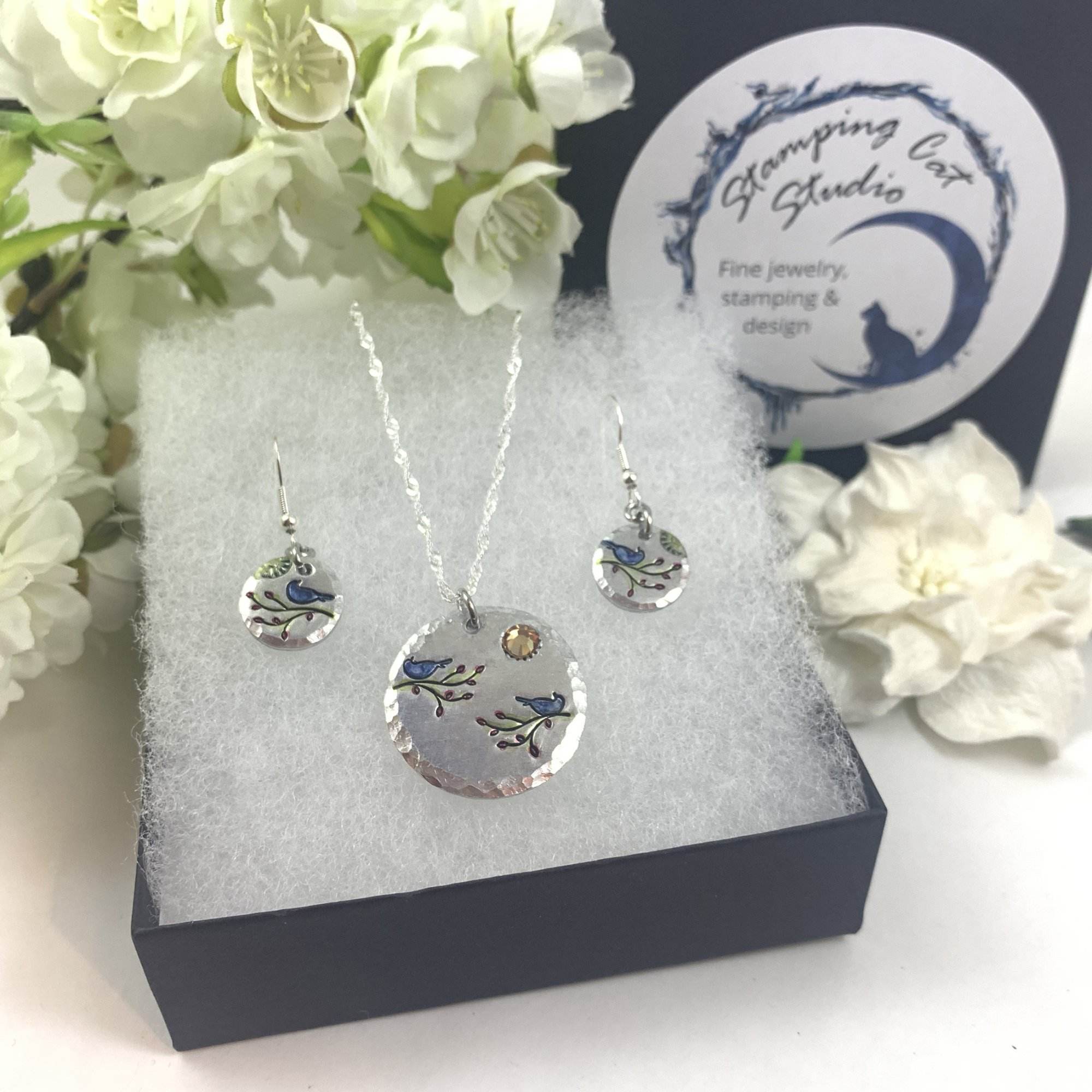 The Colorful Bird On Branch Necklace and Earring Set features round pendants with blue bird and branch designs, displayed in a black box with white padding, white flowers in the background, and a visible “Stamping Cat Studio” label. Read more at Stamping Cat Studio (stampingcatstudio.com).