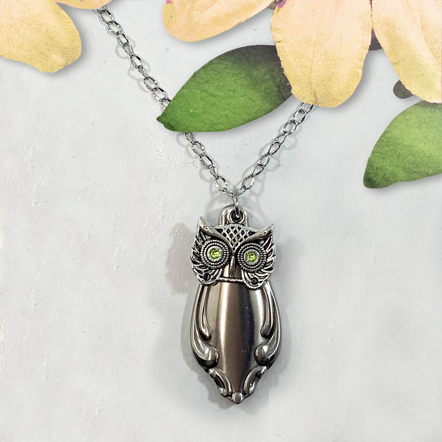 The Cute Owl Pendant Necklace features a silver owl with green gem eyes, detailed engraving for feathers and wings, and hangs from a thin chain. Displayed on a white surface with yellow and green artificial flower petals above it. Read more at Stamping Cat Studio (stampingcatstudio.com).