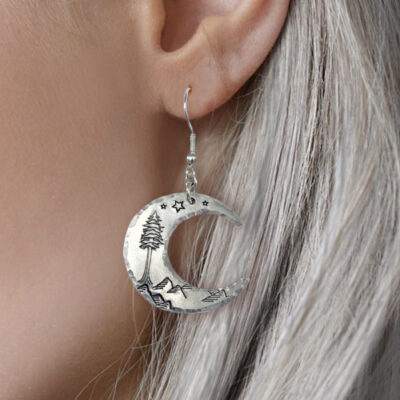 A close-up of a person with long, silver hair wearing Crescent Moon Dreamy Mountain Scene Earrings, featuring engraved stars, mountains, and a pine tree inspired by dreamy landscapes. Read more at Stamping Cat Studio (stampingcatstudio.com).