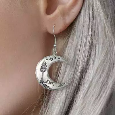 A close-up of a person with long, silver hair wearing Crescent Moon Dreamy Mountain Scene Earrings, featuring engraved stars, mountains, and a pine tree inspired by dreamy landscapes. Read more at Stamping Cat Studio (stampingcatstudio.com).