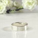 A Personalized Sterling Silver Band, 6mm with a “925” stamp inside rests on a glossy white surface, while soft white flowers and green stems in the blurred background create a delicate, minimalistic atmosphere. Read more at Stamping Cat Studio (stampingcatstudio.com).