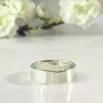 A Personalized Sterling Silver Band, 6mm with a “925” stamp inside rests on a glossy white surface, while soft white flowers and green stems in the blurred background create a delicate, minimalistic atmosphere. Read more at Stamping Cat Studio (stampingcatstudio.com).