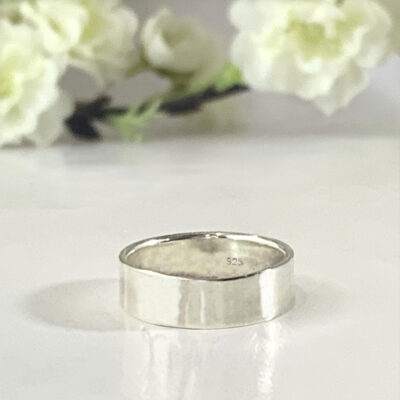 A Personalized Sterling Silver Band, 6mm with a “925” stamp inside rests on a glossy white surface, while soft white flowers and green stems in the blurred background create a delicate, minimalistic atmosphere. Read more at Stamping Cat Studio (stampingcatstudio.com).