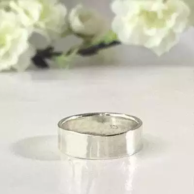 A Personalized Sterling Silver Band, 6mm with a “925” stamp inside rests on a glossy white surface, while soft white flowers and green stems in the blurred background create a delicate, minimalistic atmosphere. Read more at Stamping Cat Studio (stampingcatstudio.com).